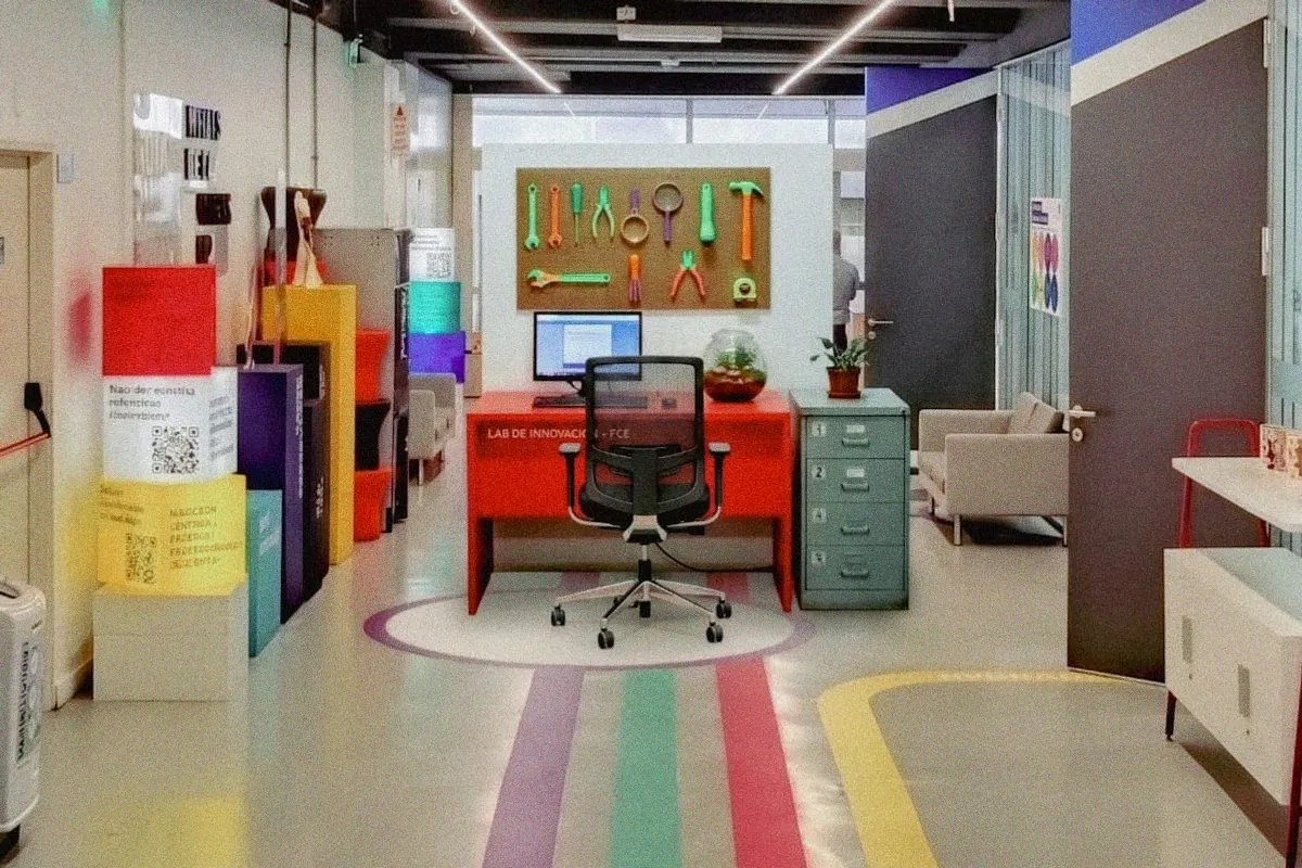 Un escritorio rojo en el pasillo de un edificio universitario, con un panel de herramientas de colores flúo detrás y un fichero de cajones numerados al costado