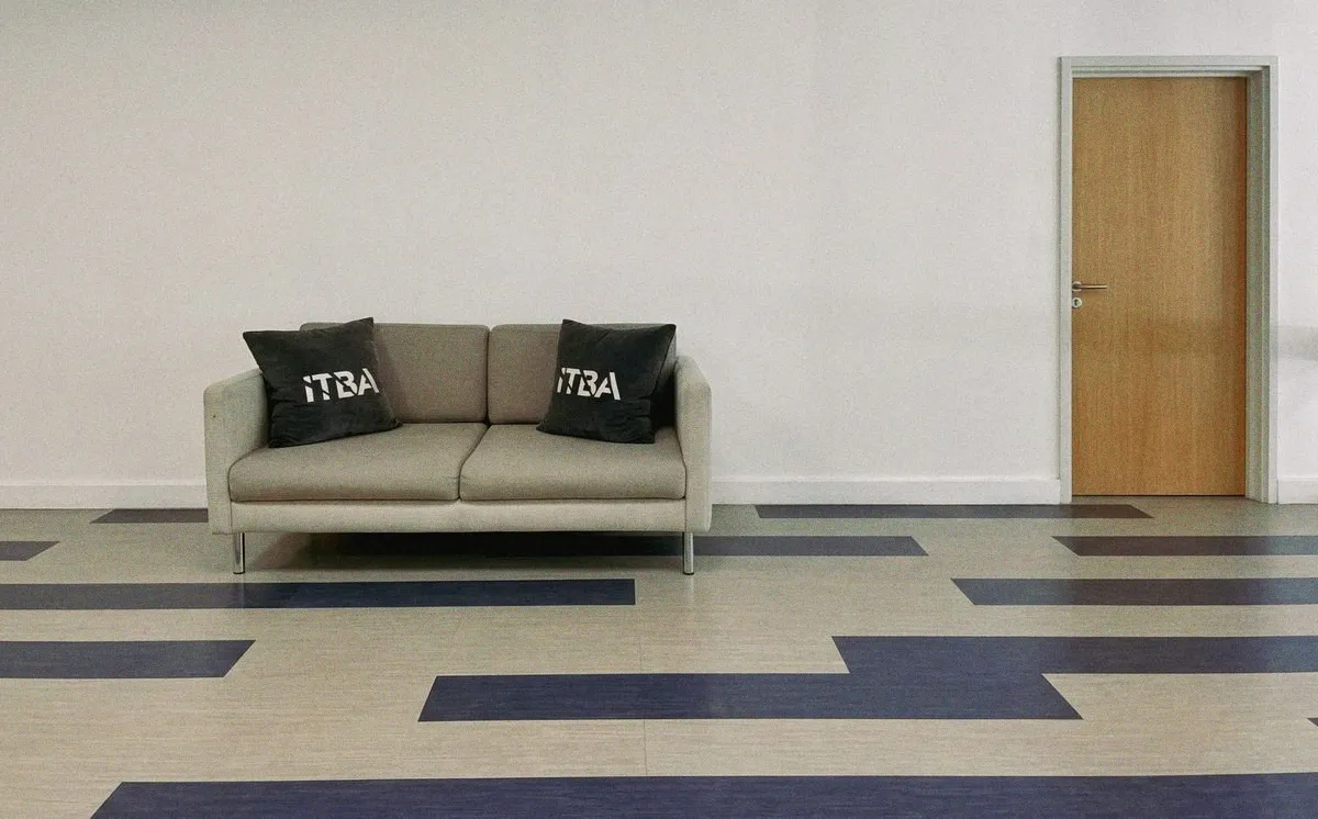 Un sillón gris vacío contra una pared blanca, con dos almohadones con el logo del ITBA, frente a una puerta cerrada de madera clara, sobre un piso vinílico con franjas azul marino