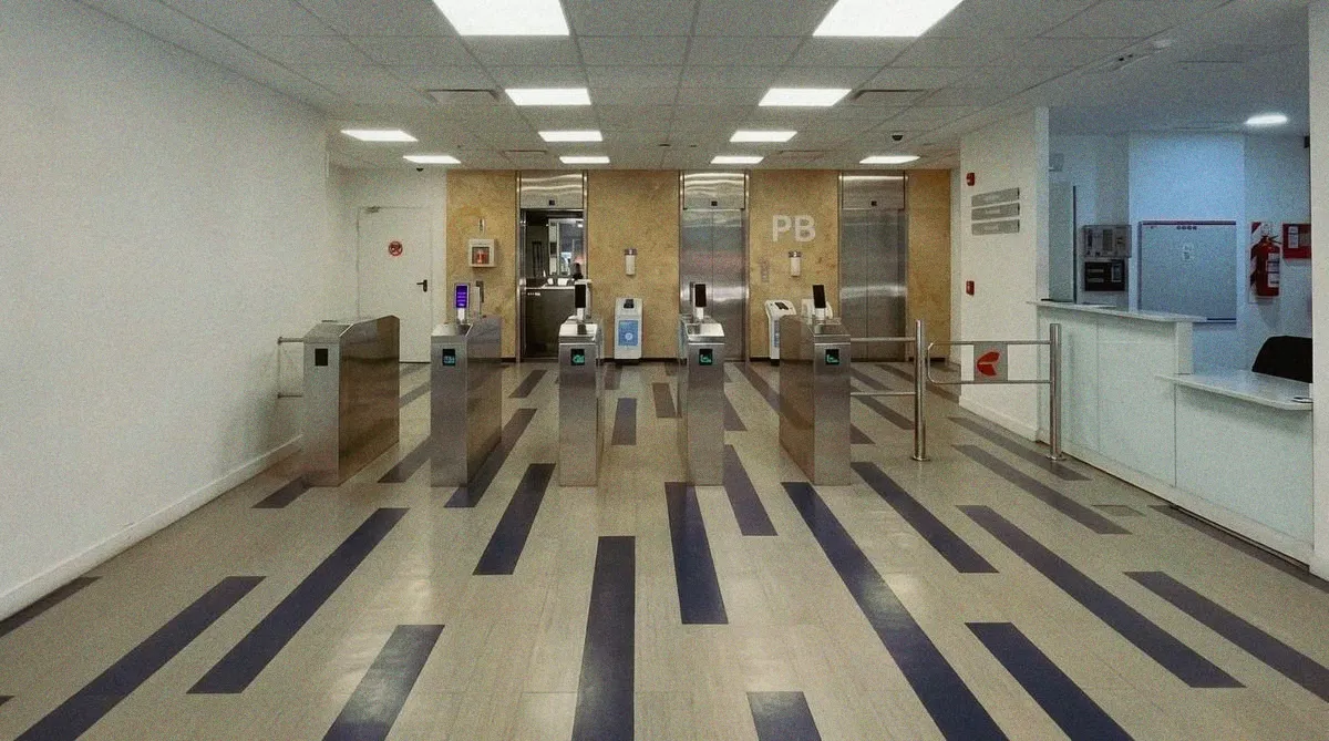 Planta baja del edificio: molinetes de acceso, tres ascensores al fondo bajo un cartel que dice PB, piso vinílico claro con franjas azules y cielorraso de paneles fluorescentes