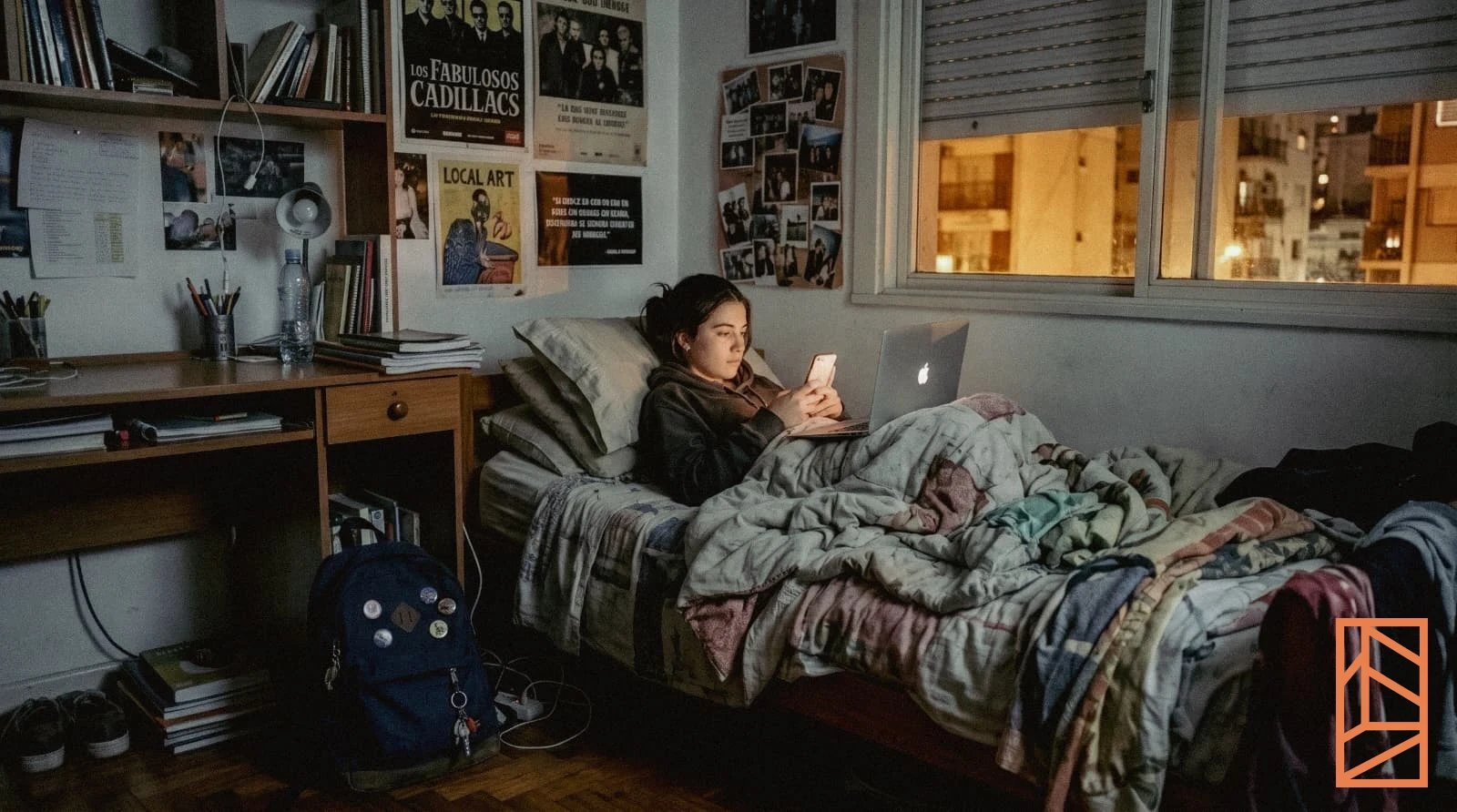 Una chica en la cama de su cuarto, de noche, con el celular en la mano y la notebook abierta. La única luz viene de las pantallas y de la ventana.